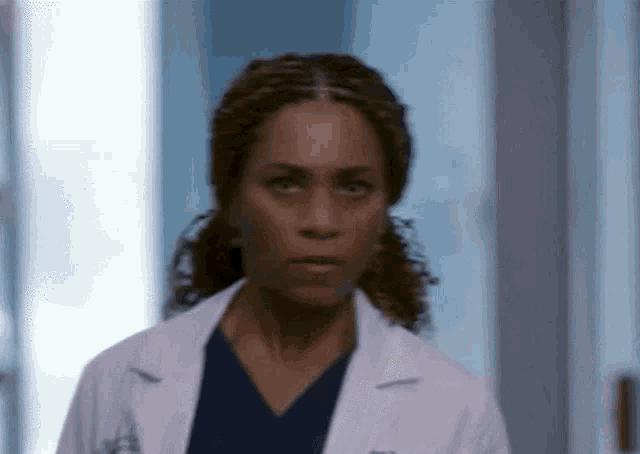 a woman in a lab coat and scrubs is looking at the camera .