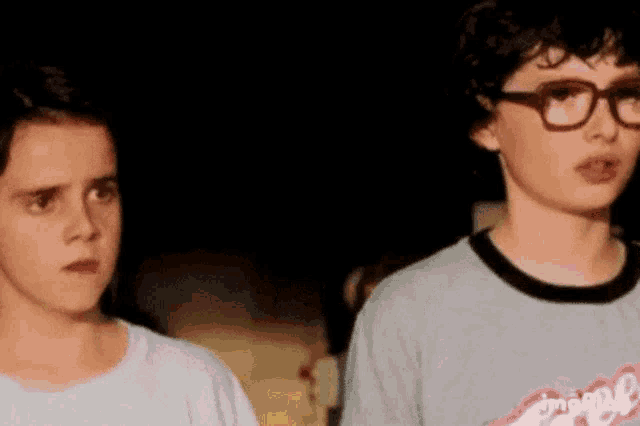 two young boys are standing next to each other in a dark room . one of the boys is wearing glasses .