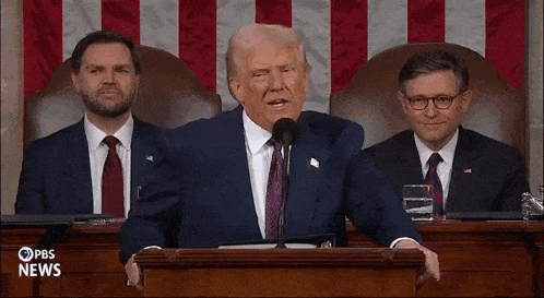 a man in a suit and tie is giving a speech in front of a microphone while two other men sit behind him .