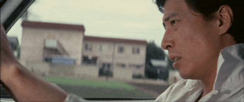 a man in a white shirt is driving a car with a building in the background