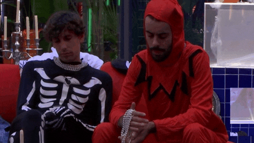 two men dressed in skeleton and devil costumes sit on a couch