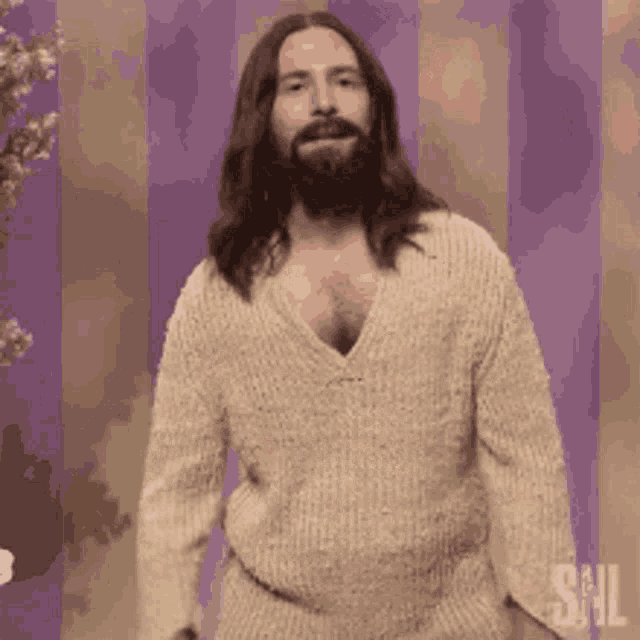 a man with long hair and a beard is standing in front of a purple wall wearing a sweater .