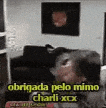 a man is sitting on a couch in a living room with the words obrigada pelo mimo charli xcx on the bottom .