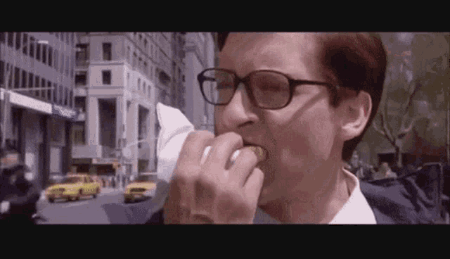 a man wearing glasses is eating a hot dog on the street .