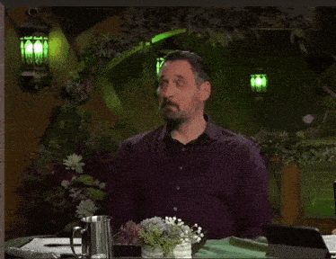 a man in a purple shirt is sitting at a table and says " nat1 on "