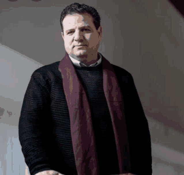 a man wearing a black sweater and a purple scarf stands in front of a white wall .