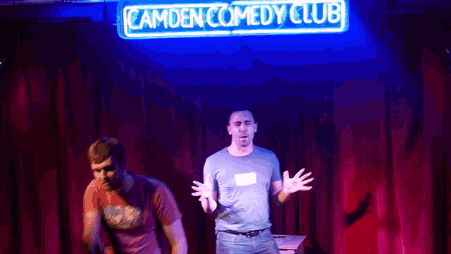 two men stand on stage under a camden comedy club neon sign