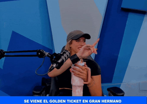 a woman holding a cup in front of a microphone with the words se viene el golden ticket en gran hermano below her