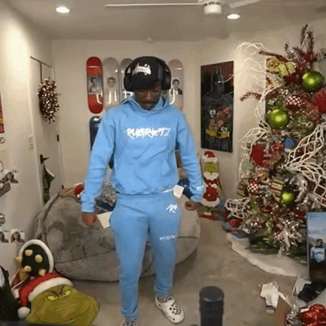 a man in a light blue hoodie and sweatpants is standing in front of a christmas tree .