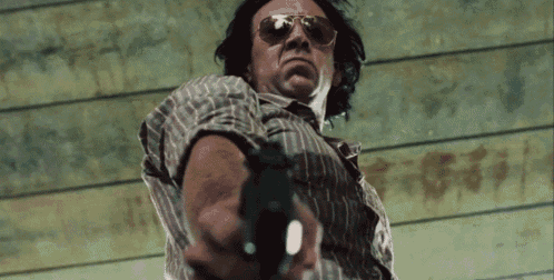 a man wearing sunglasses holds a gun in front of a wooden wall