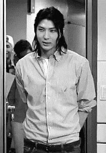 a black and white photo of a man with long hair standing in front of a door .