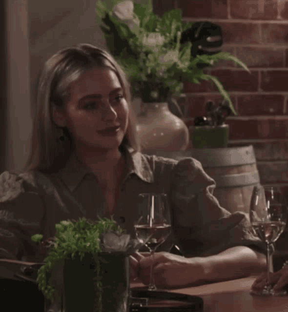a woman sits at a table with wine glasses in front of a brick wall