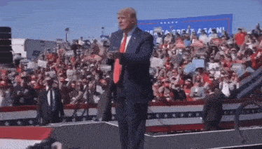 a man in a suit and tie is standing in front of a crowd with a sign behind him that says " great again "