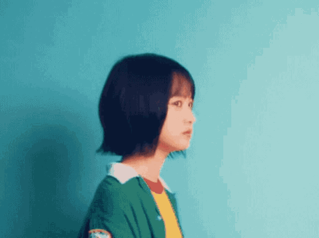 a young girl with short hair is standing in front of a blue wall wearing a green jacket and a yellow shirt .