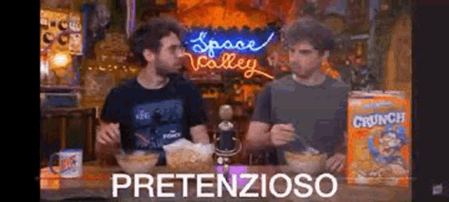 two men are sitting at a table with bowls of cereal and a box of crunch cereal .