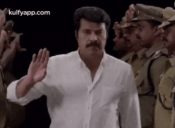 a man with a mustache is standing in front of a group of police officers and waving his hand .