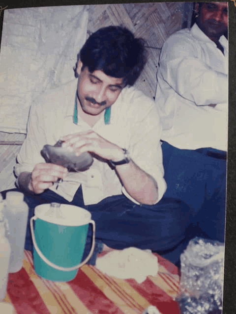 a man with a mustache is sitting on the floor with a green bucket on the table