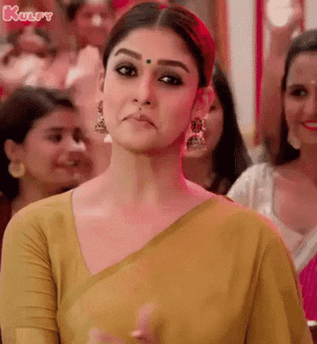 a woman in a yellow saree is standing in front of a crowd of people and making a funny face .