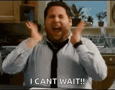 a man in a tie is standing in a kitchen with his hands in the air and says `` i can 't wait ! ''