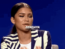 a woman in a striped jacket is sitting in front of a blue background with the word zendaya written on her face .