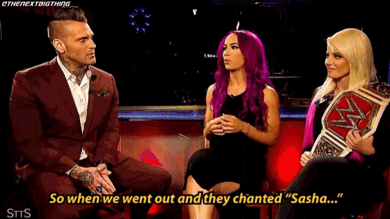 a man in a suit is sitting next to two women with purple hair and one of them is holding a wrestling championship belt .