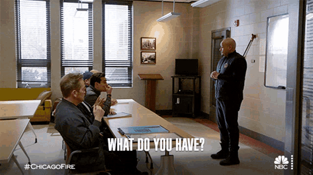 a group of men are sitting around a table with a man standing in front of them asking them what do you have