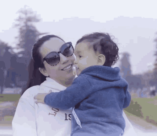 a woman wearing sunglasses holds a baby in her arms and the baby is wearing a sweater that says 19 on it