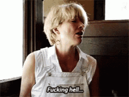 a woman wearing overalls says " fucking hell " in front of a window