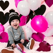 a baby wearing a shirt that says oh my is sitting in front of balloons .