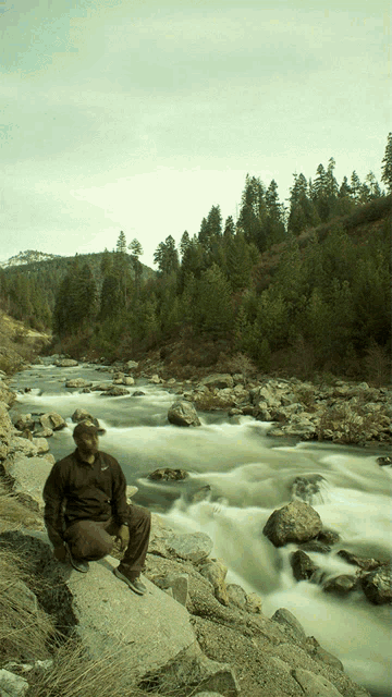 a man sits on a rock near a river with a mountain in the background