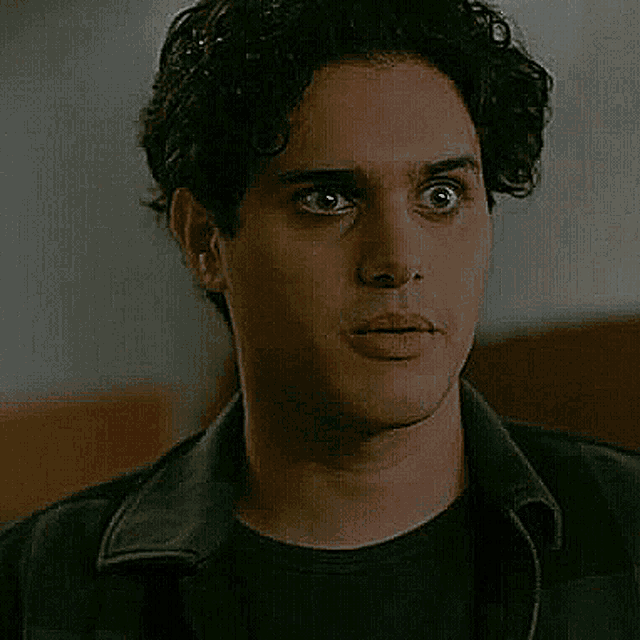 a man with curly hair is wearing a black shirt and a black jacket and looking at the camera .