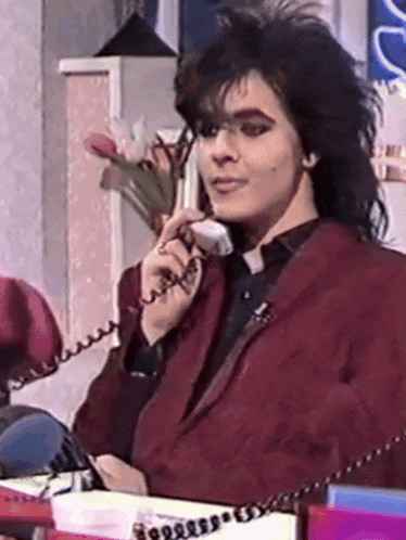 a man in a red jacket is sitting at a desk talking on a phone