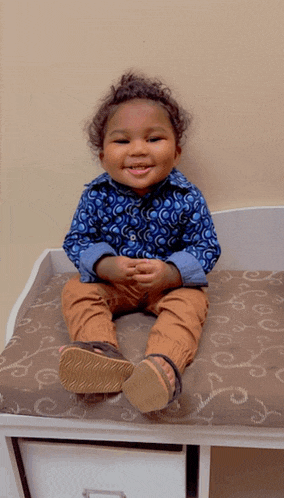 a little boy wearing a blue shirt and brown pants is sitting on a cushion