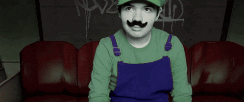 a man in a green hat and blue overalls is sitting on a couch