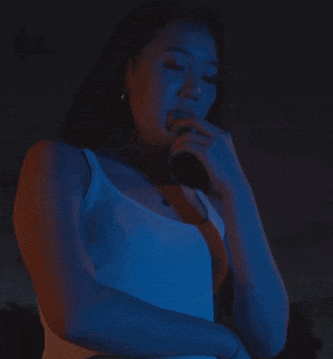 a woman in a white tank top stands in a dark room with a blue light behind her