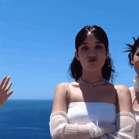 two women are standing next to each other in front of the ocean . one of the women is wearing a necklace and gloves .