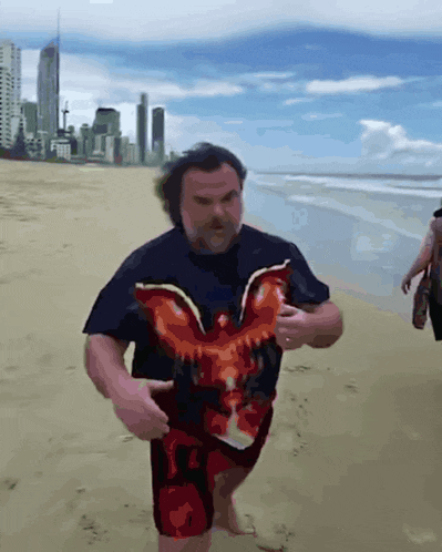 a man in a black shirt with an eagle on it is walking on the beach