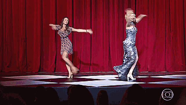 two women are dancing on a stage with red curtains behind them