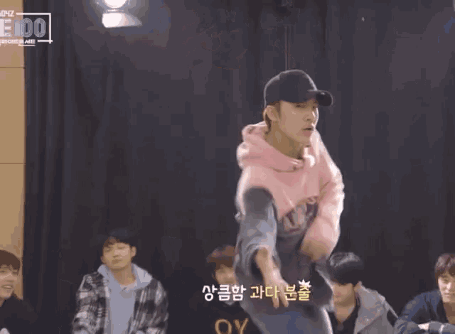 a man wearing a pink hoodie and a black hat is dancing in front of a sign that says " e100 "