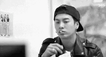 a black and white photo of a young man wearing a baseball cap and eating a sandwich .