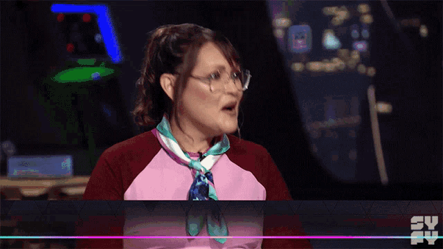 a woman wearing glasses and a pink shirt is sitting in front of a tv screen that says syfy