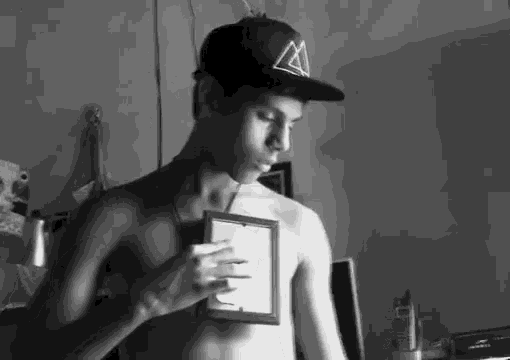 a shirtless man wearing a hat holds a picture frame in his hand