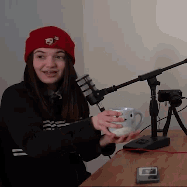 a woman wearing a red beanie is holding a coffee mug in front of a microphone