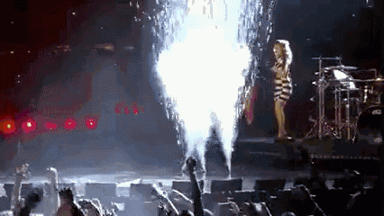 a woman is standing on a stage in front of a crowd with a fireworks display in the background