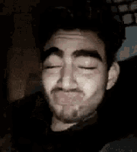 a man with a beard is making a funny face with his eyes closed in a dark room .