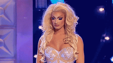 a drag queen is standing on a stage wearing a white and gold dress and a wig .