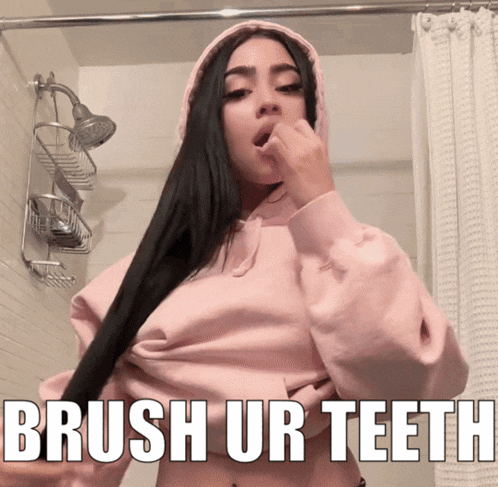 a woman brushes her teeth in a bathroom while wearing a pink hoodie