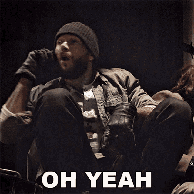 a man in a beanie sits in a dark room with the words oh yeah above him
