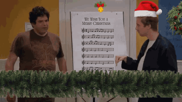 a man wearing a santa hat stands next to a sheet of music that says we wish you a merry christmas