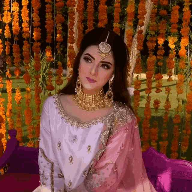 a woman in a white and pink dress is sitting in front of a wall of orange flowers